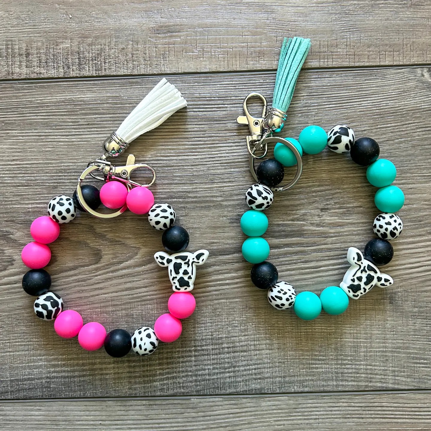 Two cow-themed silicone bead wristlet keychains in hot pink and teal with cow print beads and tassels
