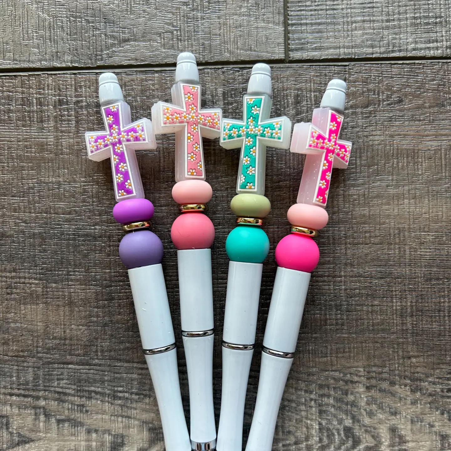 Four colorful beaded pens with daisy cross toppers in purple, pink, teal, and hot pink